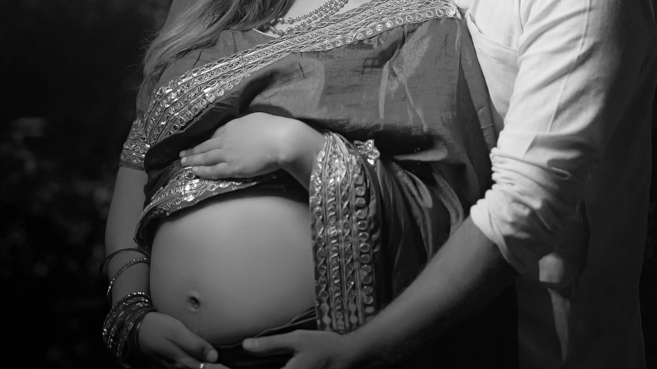 Close-up black and white photography of an Indian maternity photoshoot by Indo Frames, showing the husband's and wife's hands cradling her baby bump adorned with traditional bangles.
