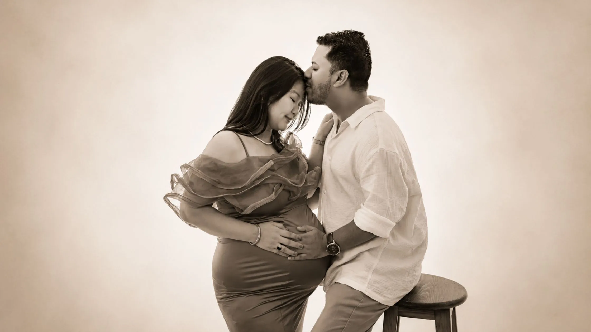 A timeless black and white couple portrait from an indoor indian maternity photoshoot collection by Indo frames, showing a husband kissing his pregnant wife's forehead.
