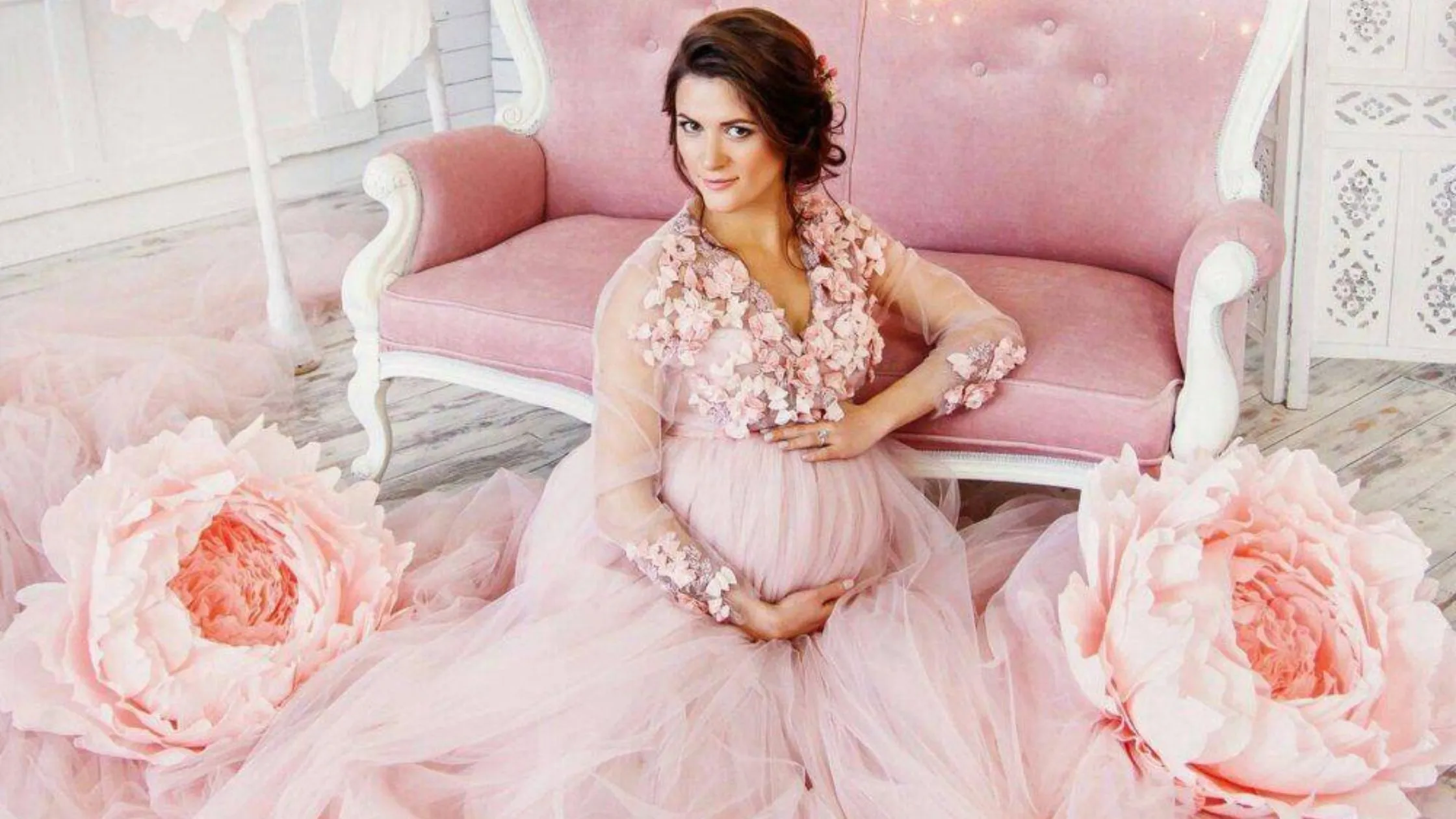 An expectant mother sitting elegantly on the floor in front of a pink velvet sofa for her maternity photoshoot poses. She is wearing a blush pink tulle dress with 3D floral details, surrounded by giant decorative paper peonies, styled by Indo frames.