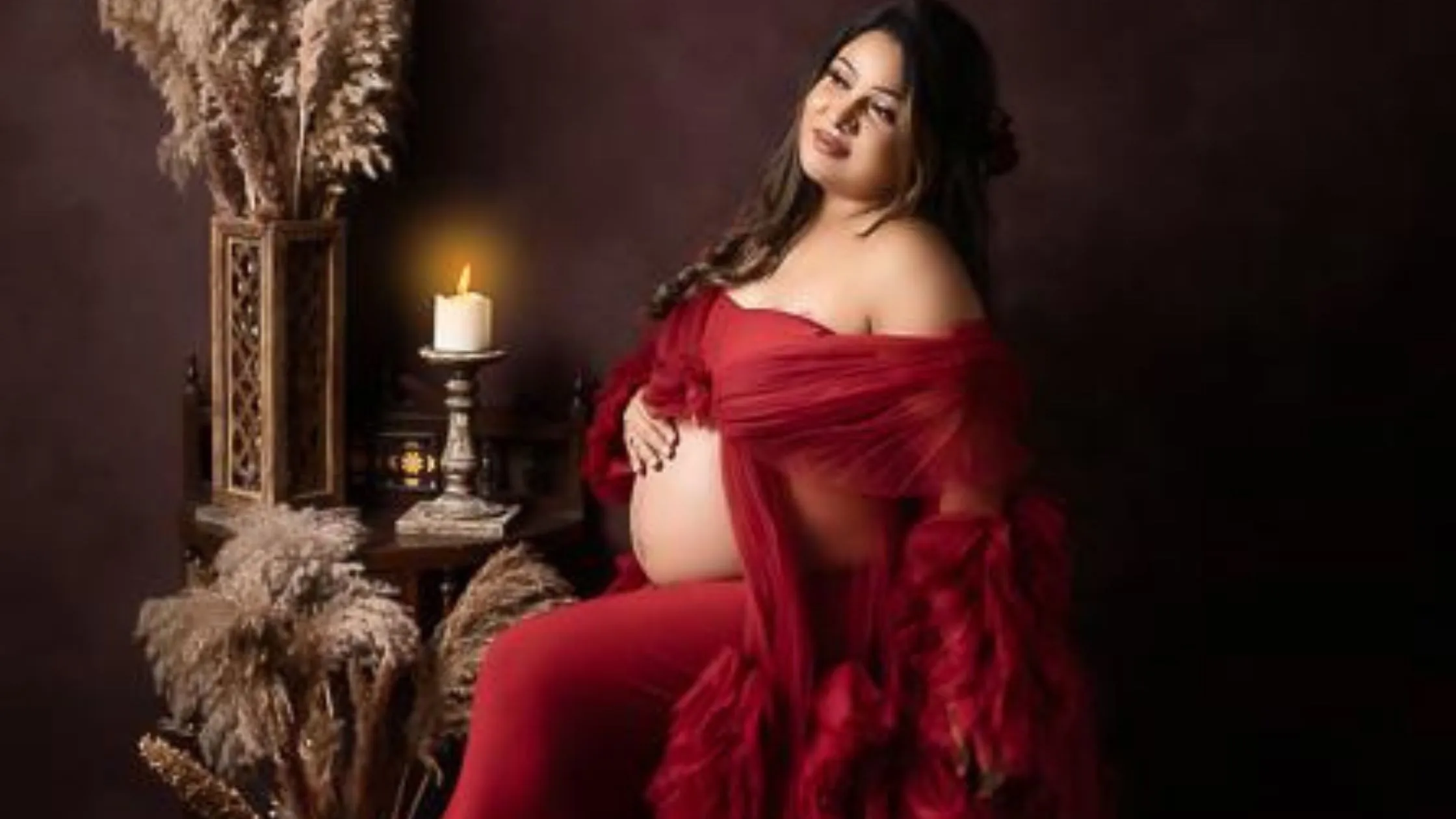 A glamorous fine art portrait showcasing maternity photoshoot poses by Indo frames. A pregnant woman in a flowing deep red off-the-shoulder tulle gown cradles her baby bump in a dark, moody studio setting illuminated by a single candle.