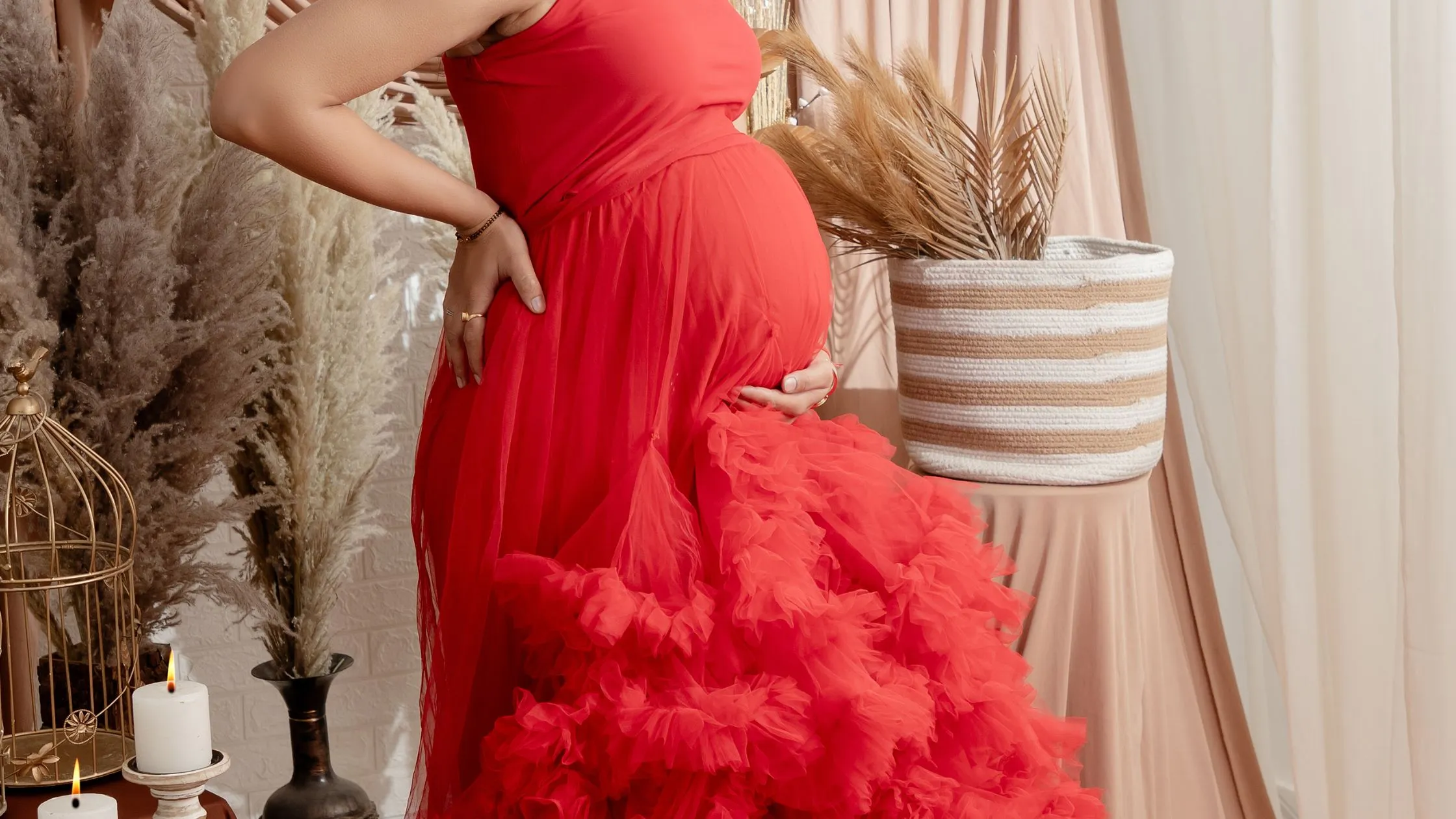 A vibrant indoor indian maternity photoshoot by Indo frames featuring an expecting mother in a luxury red ruffled gown inside a boho-themed studio setup.